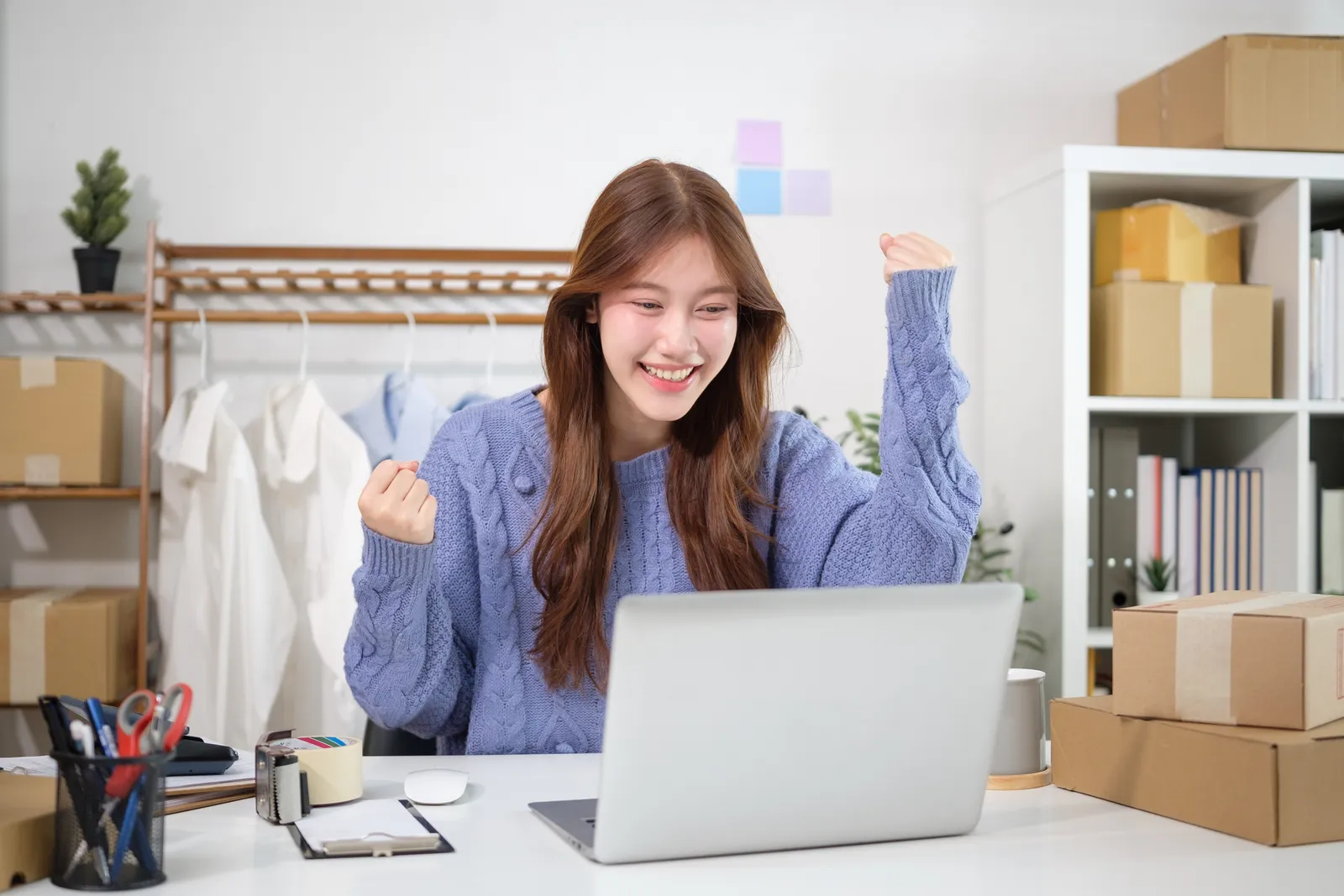 Online seller celebrating an order at her laptop beside stacked boxes