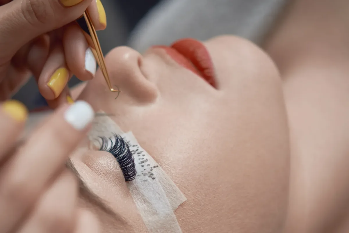 A lash technician applying eyelash extensions to a client reclined in a salon chair under a ring light