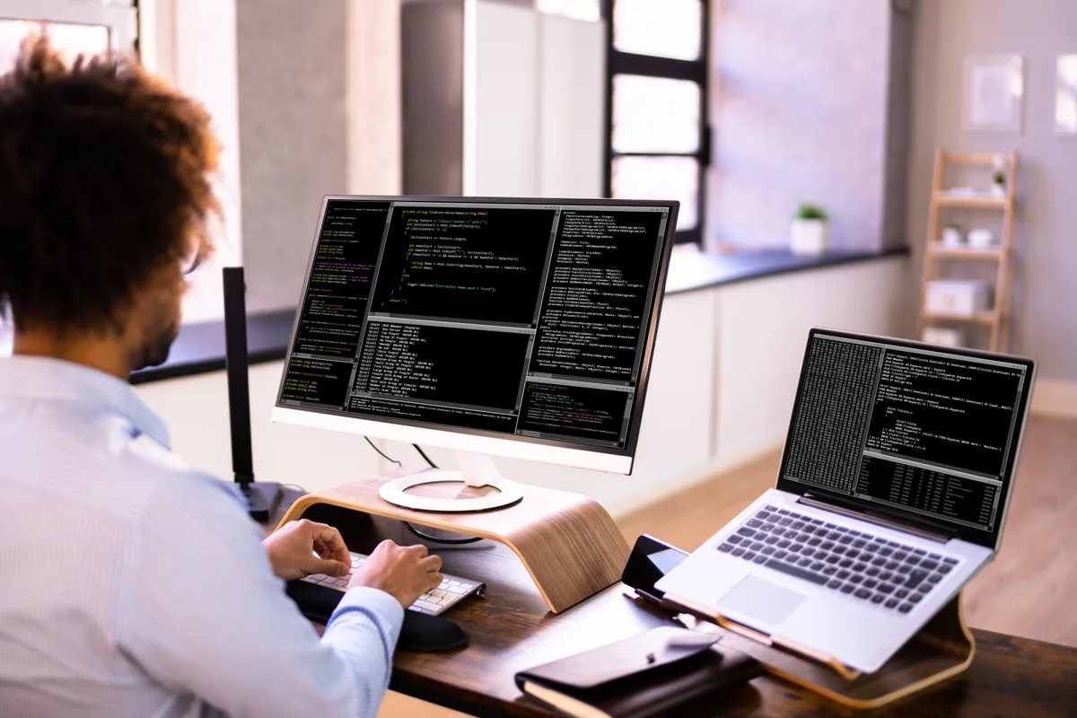 A web developer reviewing site monitoring dashboards and code on dual monitors during routine maintenance