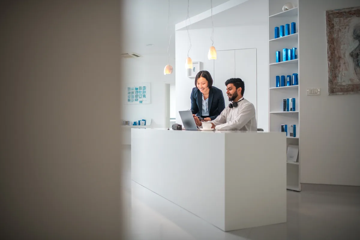 A dentist and practice manager reviewing the new-patient appointment schedule on a computer at a modern dental clinic front desk