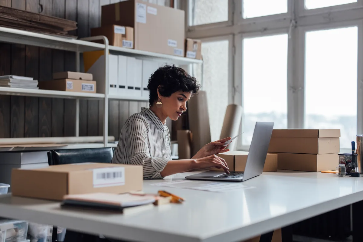 Shopify store owner reviewing product listings and search analytics on a laptop at a packing bench