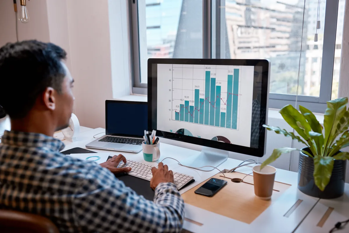 A digital marketing analyst studying a Google Ads cost-per-lead trend line on a desktop monitor while taking notes