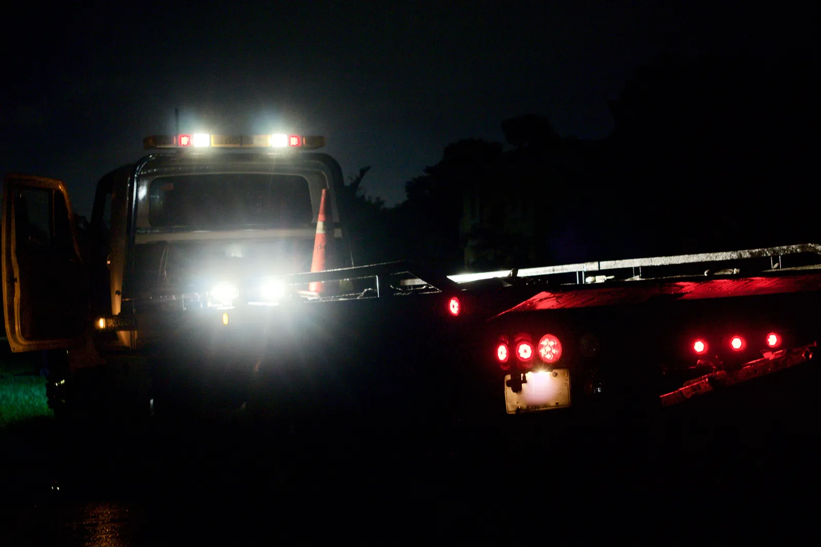 Flatbed wrecker parked at night with its work lights and amber beacons lit and a traffic cone set out beside the deck