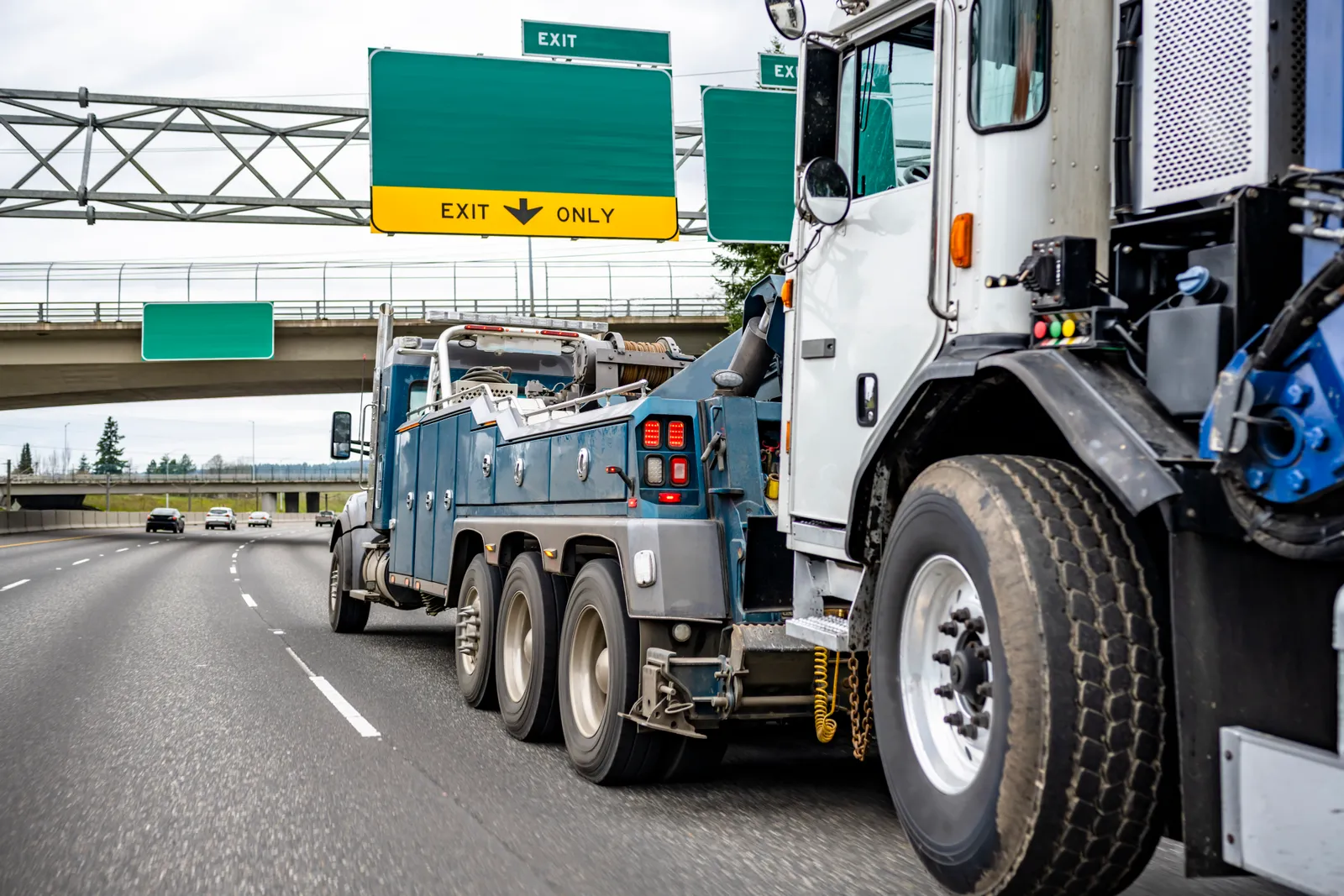 Heavy-duty wrecker towing a disabled semi tractor down a multi-lane highway under an exit sign
