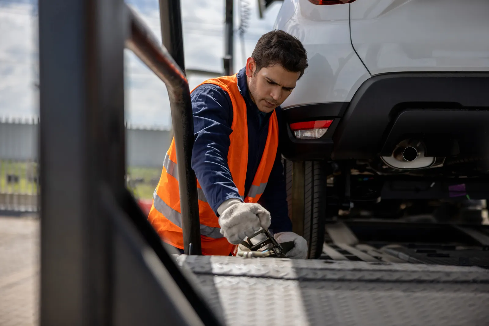 Tow operator in a hi-vis vest ratcheting a wheel strap under a white car chained down on the flatbed deck