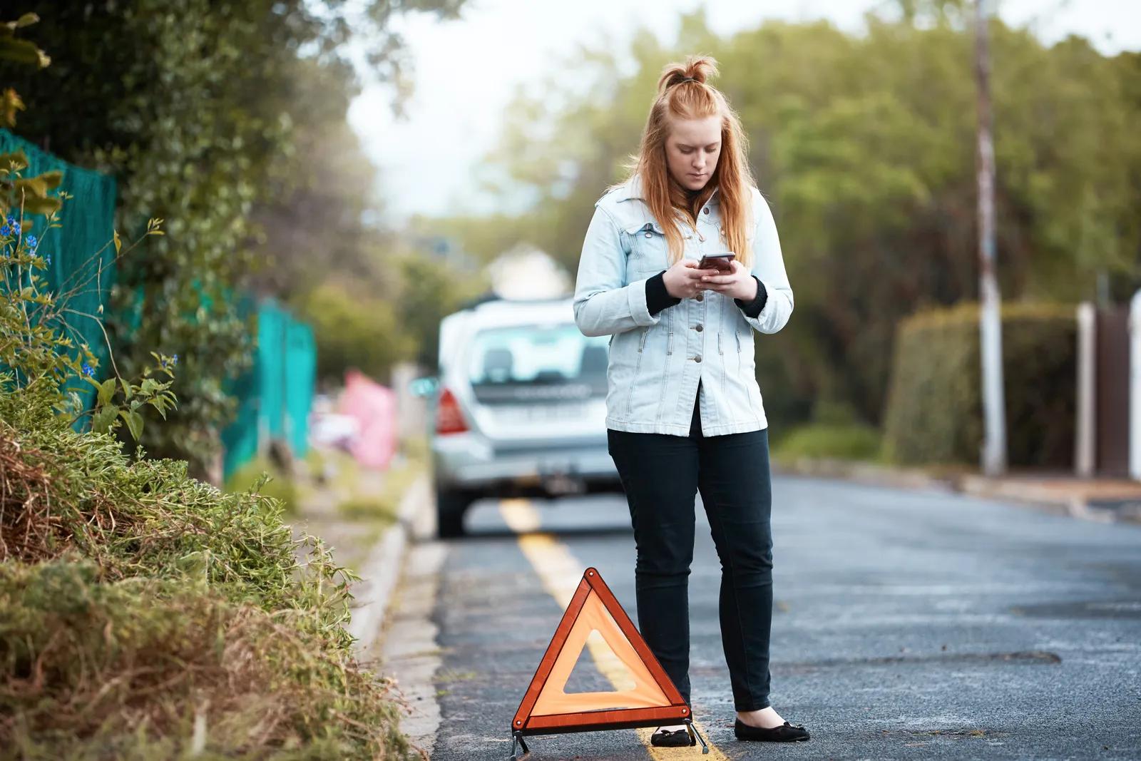 Driver standing behind her broken-down car with a reflective warning triangle set out, phoning for a tow