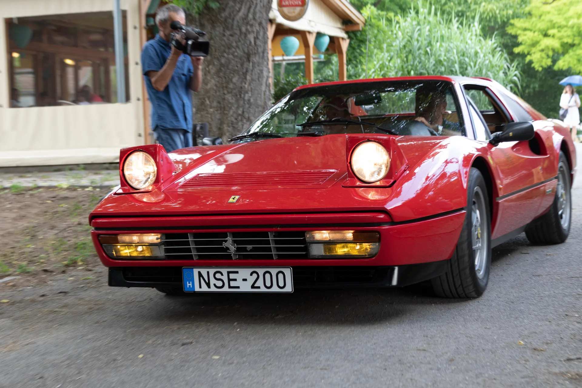 Red Ferrari 328 GTS with pop-up headlights raised, photographed from the front three-quarter on a tree-lined street