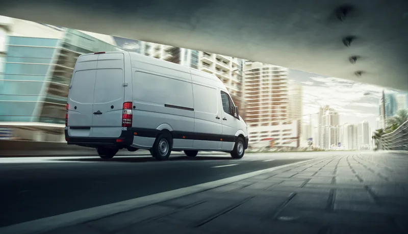 A white cargo van driving through the city on a work route