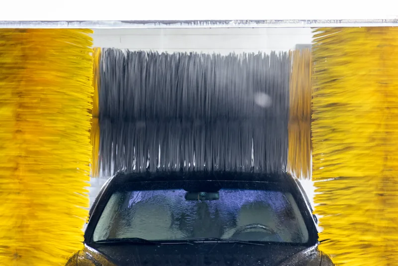 A dark car rolls under spinning yellow and gray soft-cloth brushes in a bright wash tunnel