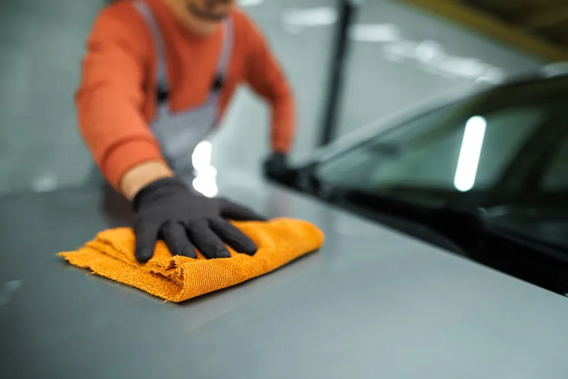 A gloved hand wiping a car hood dry with an orange microfiber towel