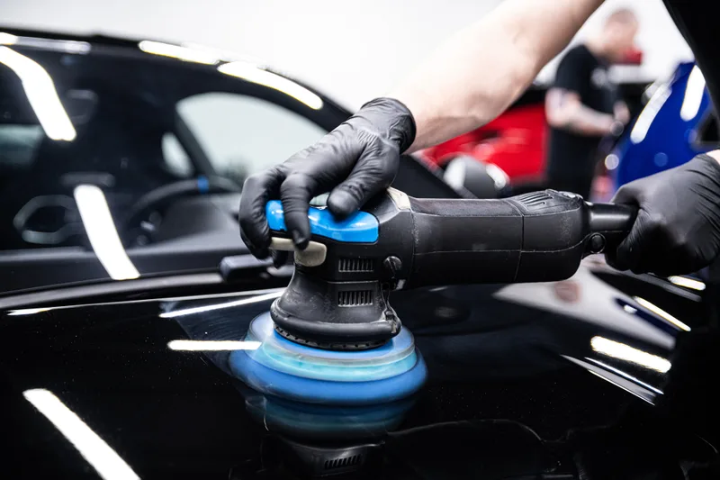 Detailer guiding a polisher with a blue foam pad across black paint under studio lights