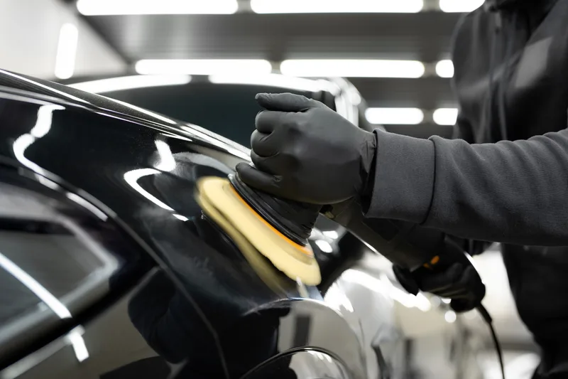 Detailer in black gloves running an orbital polisher across the glossy black paint of a car in a lit studio