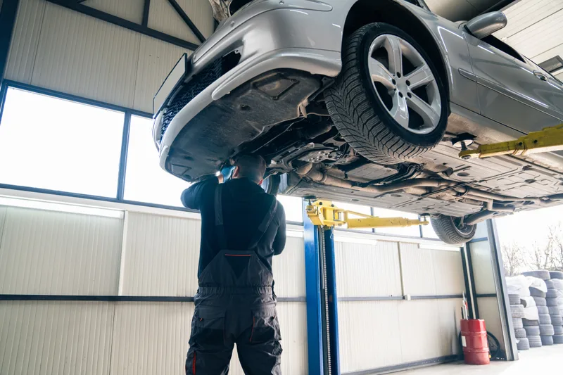 Technician working under a silver car raised on a lift in a bright bay, winter tires on the wheels and bagged stored tires stacked by the door