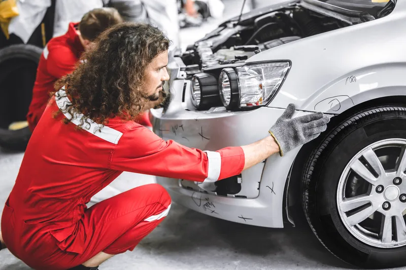 Technician kneeling to check the damaged front bumper and headlamp area of a silver car