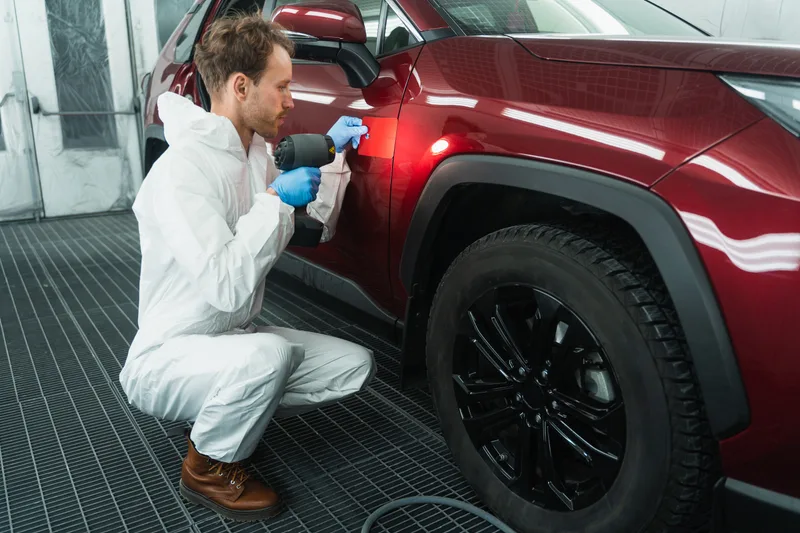 Refinish technician in white coveralls crouching beside a red SUV in a paint booth, checking the color match with a handheld lamp