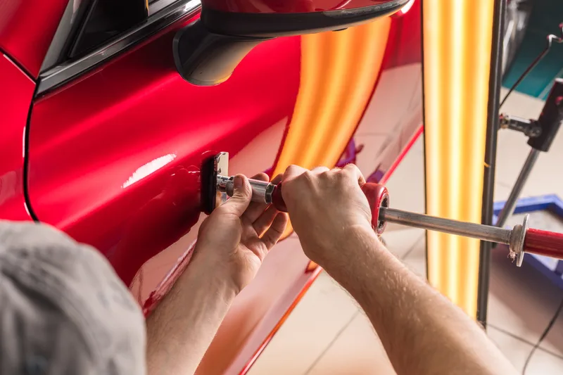 Technician's hands using a pull tool to lift a dent from a red door under a striped inspection light