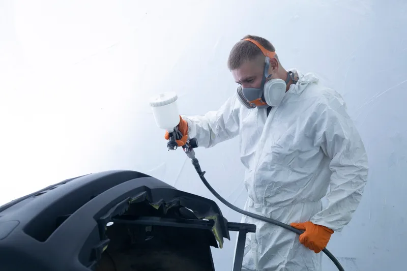 Painter in a respirator and white coveralls spraying a bumper cover in a clean paint booth