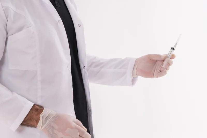A clinician in a white coat and exam gloves holding a prepared syringe at their side