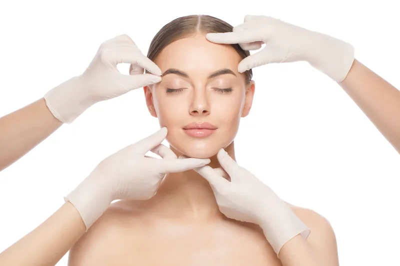Four gloved clinicians' hands gently framing a woman's relaxed face during a facial assessment