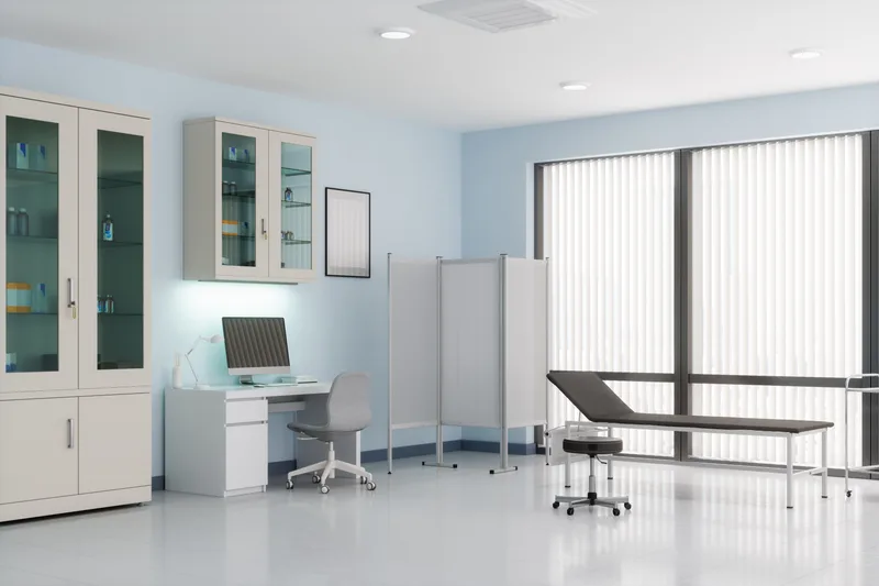 A bright private treatment room with a padded table, supply cabinets, and a privacy screen
