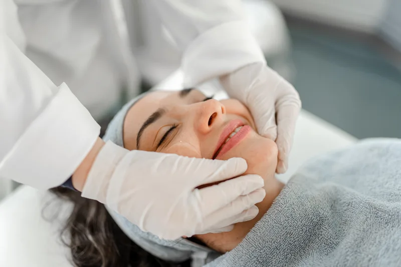 Gloved hands performing a facial treatment on a relaxed patient wrapped in a soft grey towel