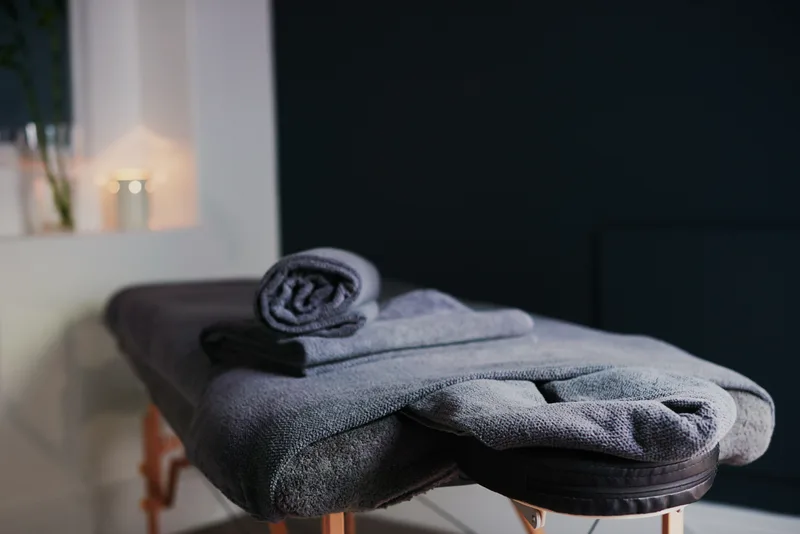 Rolled slate-grey towels stacked on a massage table in a dim, calm room with a lit candle on the sill