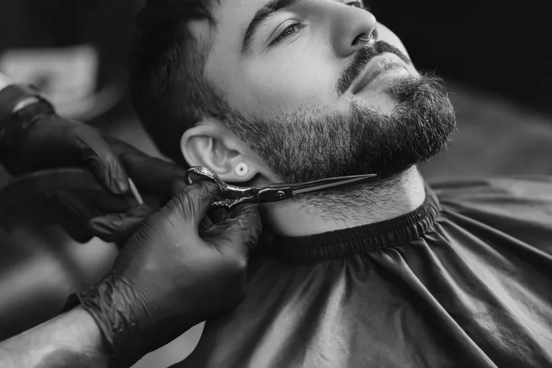 Black and white close-up of a barber shaping a full beard with scissors