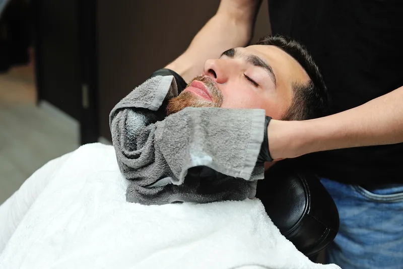 Client reclined in a barber chair with a warm towel wrapped around his jaw before a shave