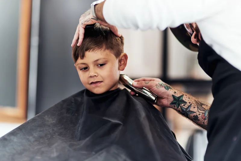 Young boy in a black cape sitting still while a barber trims the side of his hair with clippers