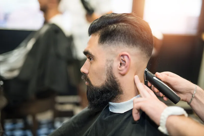 Barber blending a skin fade above the ear of a bearded client with a trimmer