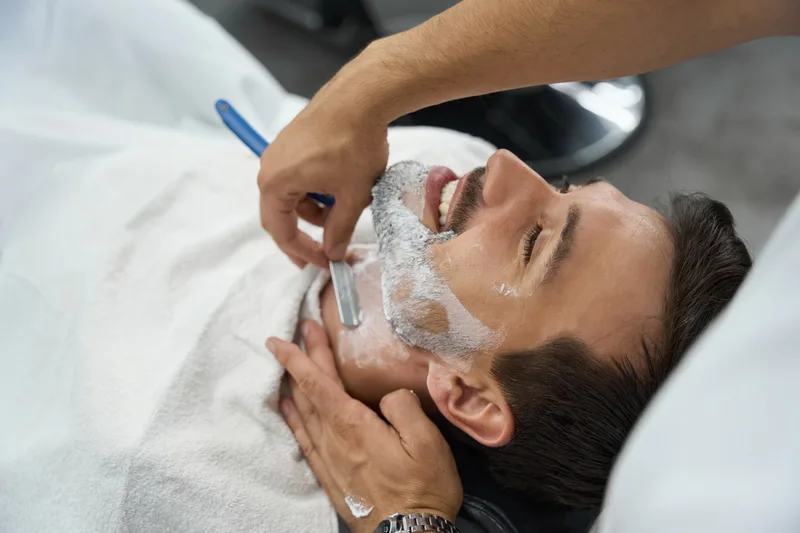 Barber shaving a lathered cheek with a straight razor while the client rests on a white towel