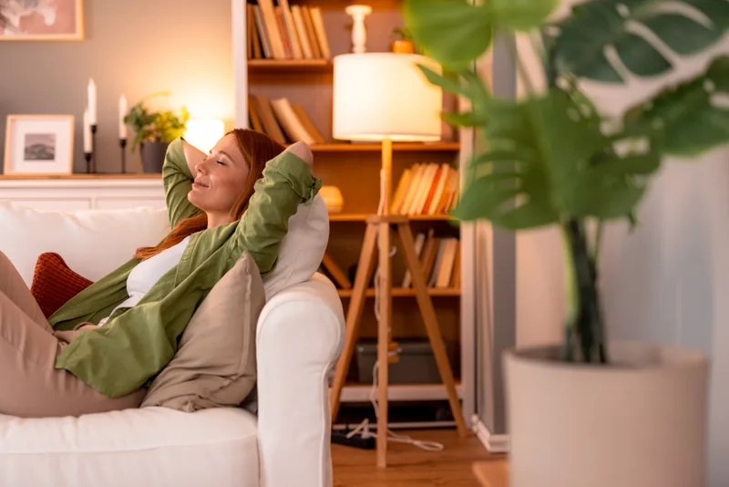 A woman resting on a sofa at home in the evening, eyes closed, hands behind her head