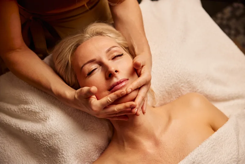 An esthetician's hands cradling a relaxed client's jaw during the massage part of a facial, client wrapped in a soft white towel
