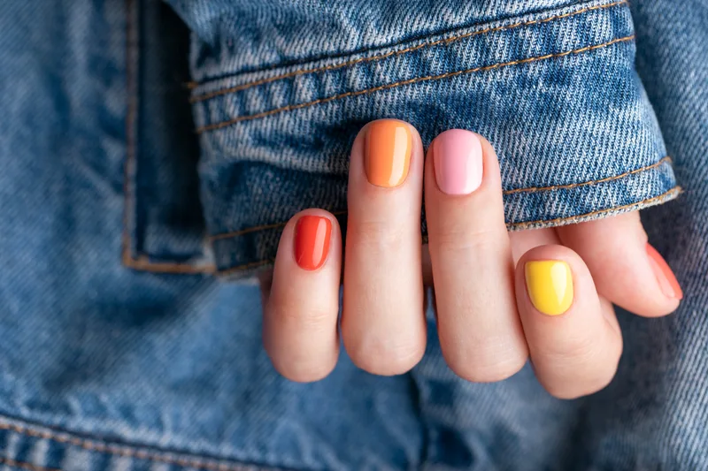 A hand with playful orange, pink, and yellow nails tucked into the pocket of a denim jacket