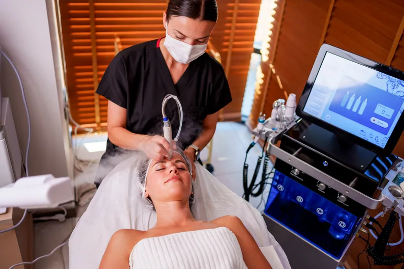An esthetician in a mask giving a facial treatment beside a professional skincare machine in a warm-lit room