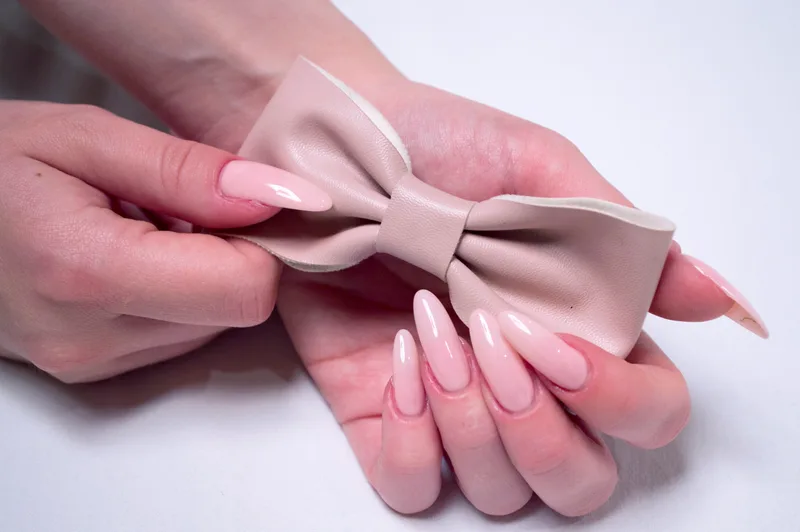Hands with glossy pink almond nails holding a small blush leather bow