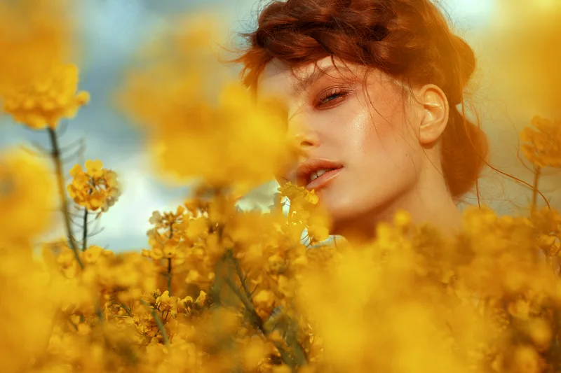 Copper-red hair pinned softly above a woman's sunlit face, glimpsed through a haze of yellow wildflowers