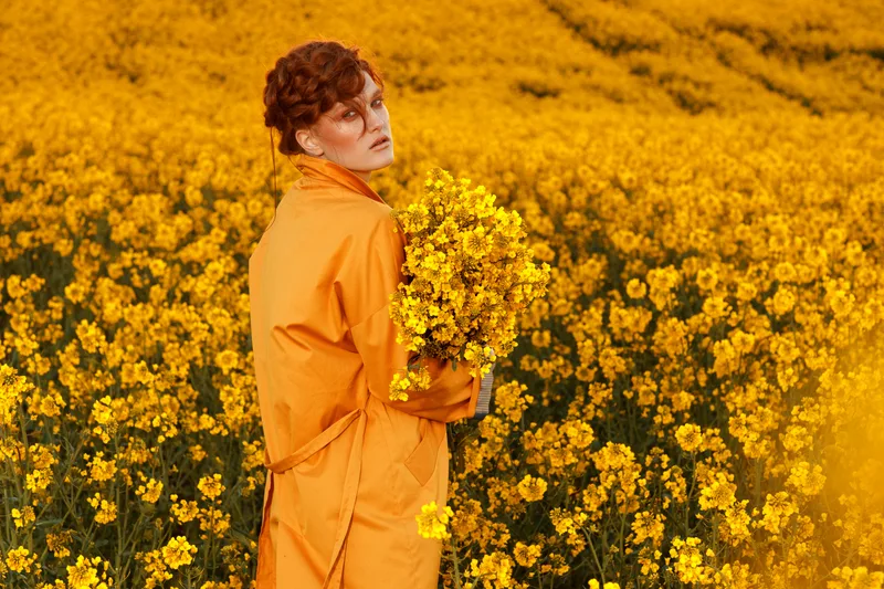 Woman with a loosely braided red updo, wearing an amber coat and holding yellow blossoms in a golden flower field
