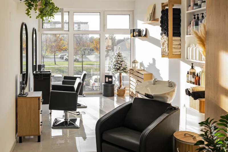 Sunlit wash corner of the salon: a cream basin chair beside shelves of folded towels and rows of haircare bottles