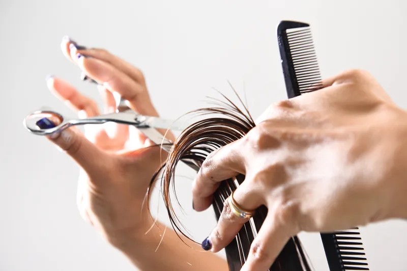 Stylist's hands mid-cut, holding a curl of brown hair between fingers with scissors and a black carbon comb