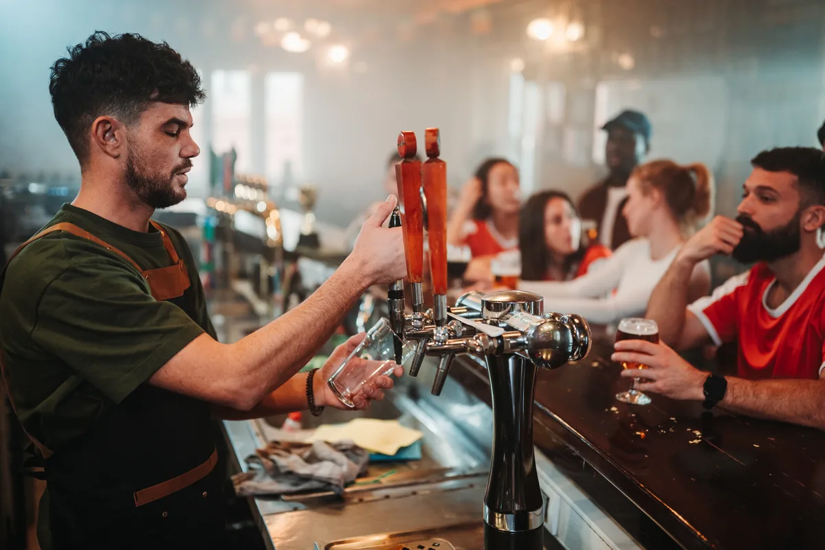 A busy craft brewery taproom with guests at the bar while a bartender pours beer from a tap