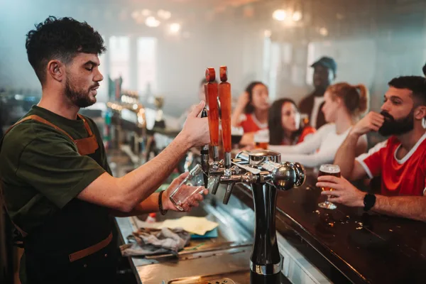 A busy craft brewery taproom with guests at the bar while a bartender pours beer from a tap