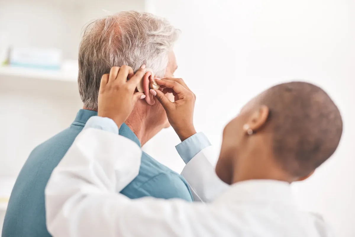 Audiologist fitting a hearing aid behind a seated older patient's ear during an appointment