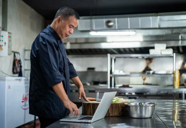 A catering company owner reviewing event bookings and a marketing proposal on a laptop in a commercial kitchen