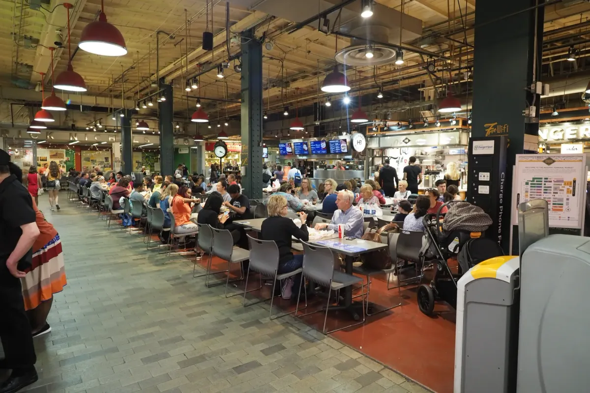 Guests seated at communal tables inside a busy food hall with lit vendor stalls lining the concourse behind them