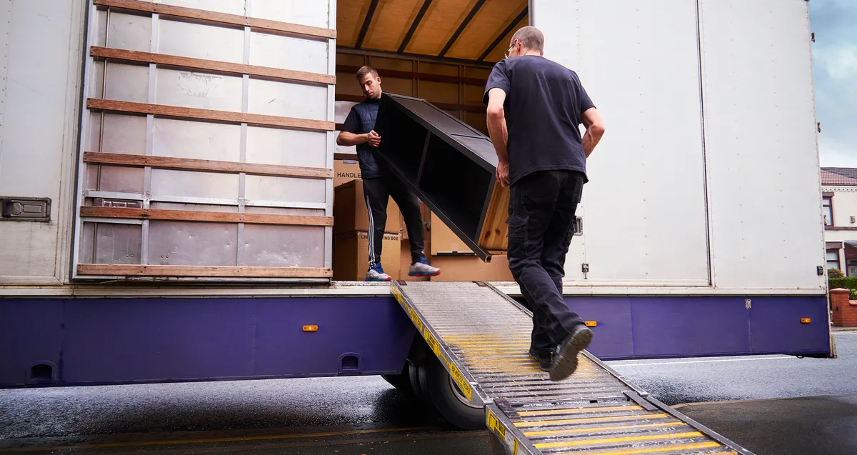 Two uniformed movers carrying furniture into a moving truck parked outside a suburban home