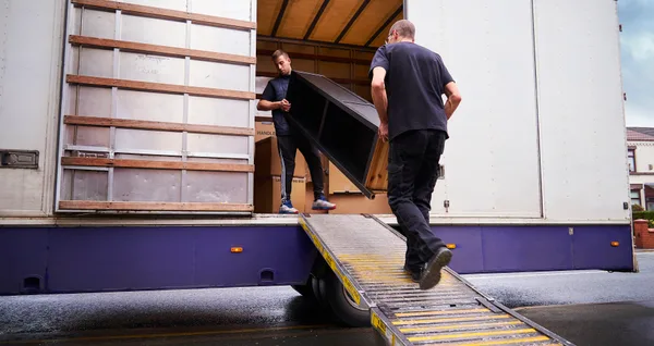 Two uniformed movers carrying furniture into a moving truck parked outside a suburban home