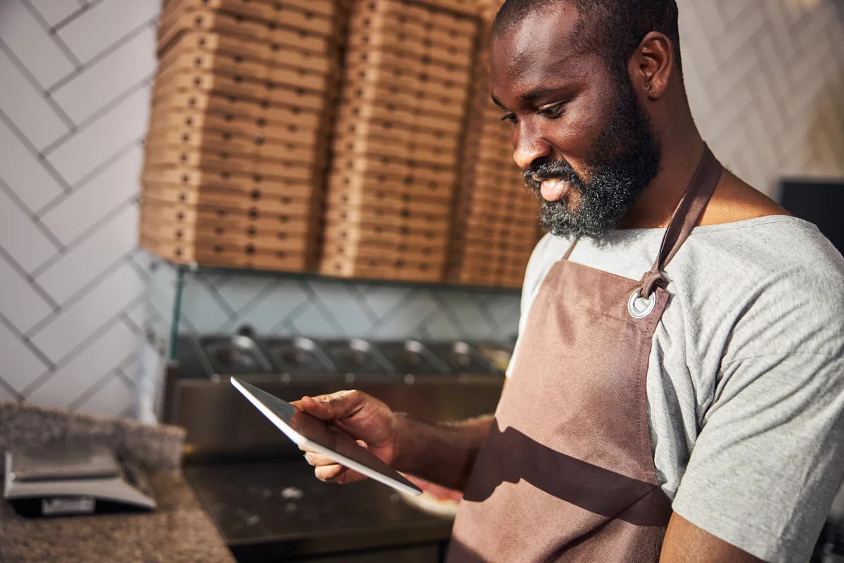 Pizzeria owner reviewing online orders on a tablet at the counter of a busy pizza shop