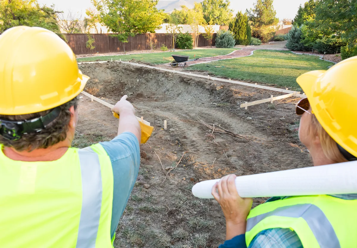 A pool builder and a homeowner reviewing inground pool design plans beside a landscaped backyard