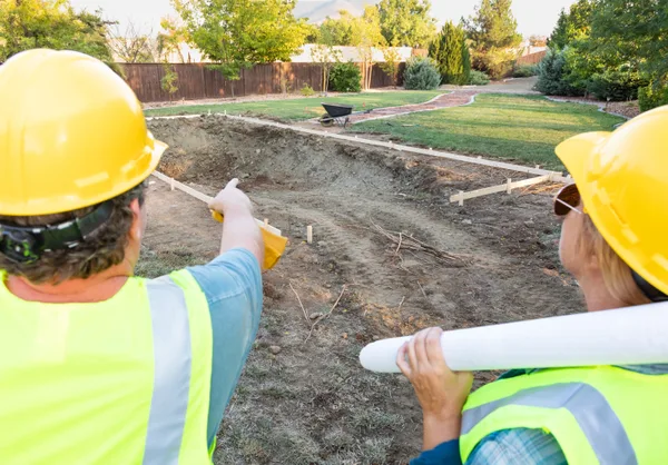 A pool builder and a homeowner reviewing inground pool design plans beside a landscaped backyard